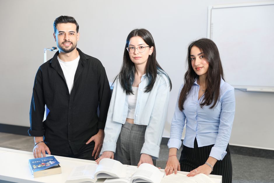 Three young professionals standing together in a modern office with books and a whiteboard.